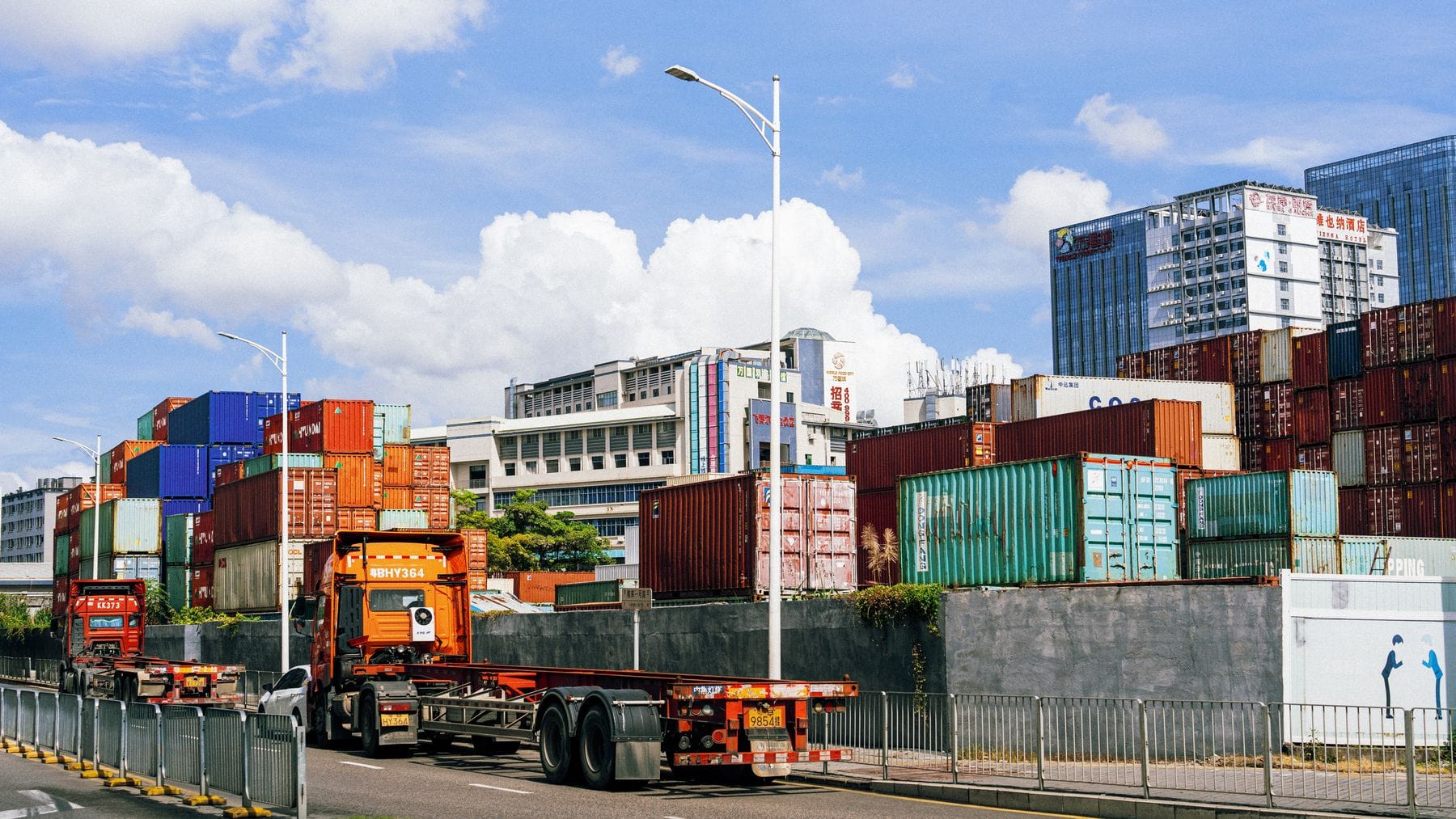 Truck hauling a shipping container past stacked cargo containers at a logistics yard
